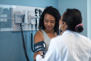 A young black woman is at a routine medical appointment. Her healthcare provider is an ethnic woman. The patient is sitting on an examination table in a clinic. The doctor is checking the patient's blood pressure. The patient is smiling while looking down at her arm. Birth control pill and cancer risk - myth or fact?